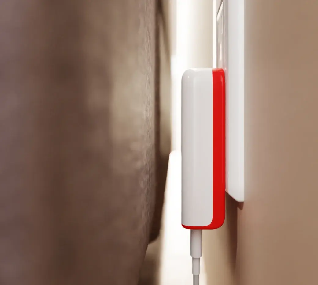 The PlugBug sitting nearly flush in a wall outlet in the narrow gap behind a sofa, its cable running down the wall